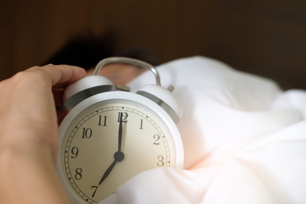 relógio despertador com uma mão segurando embaixo dos lençóis distúrbios do sono